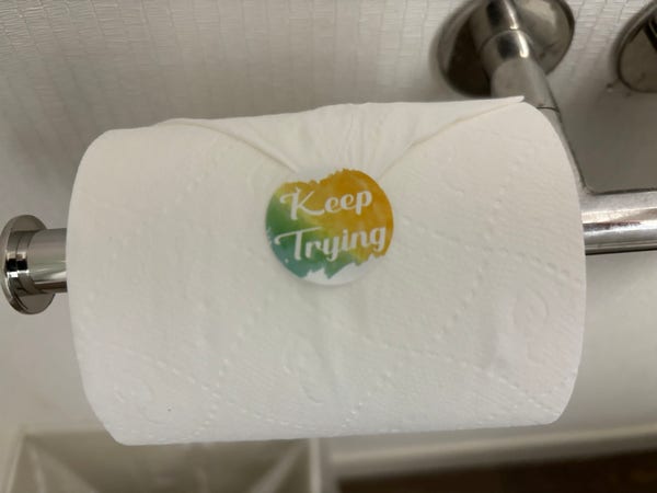 A roll of toilet paper, hung on a dispenser, with a sticker reading “Keep Trying.”