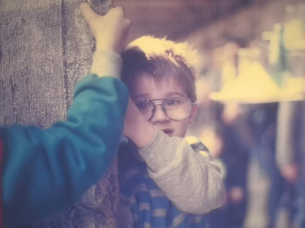 Throwback photo of me as a young boy in the mid-1980s, standing against a rough textured wall. I'm wearing a cozy winter outfit, partially hiding my face behind my arm. My glasses slightly magnify my eyes as I gaze thoughtfully towards something outside the frame. The image has a soft focus and a vintage color palette that gives it a nostalgic feel.