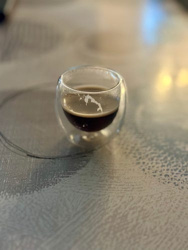 Un verre transparent à double paroi contenant un liquide foncé, peut-être du café, sur une table à motifs. L'accent est mis sur le verre et l'arrière-plan est flou.