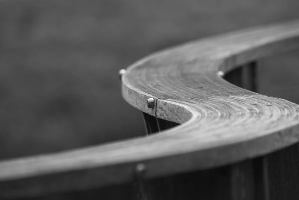 A black and white photo of a serpentine shaped deck railing made from hardwood laminated on edge and held together with stainless steel hardware drilled through horizontally.  Only a narrow slice of the view is in focus giving it an ethereal presence.