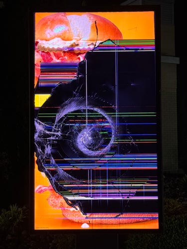 A damaged digital display at a McDonald’s drive-thru, showing a partial burger at the top with significant glitched output below. A spiral in the golden ratio originates from a likely point of impact (from a rock or something similar, perhaps).