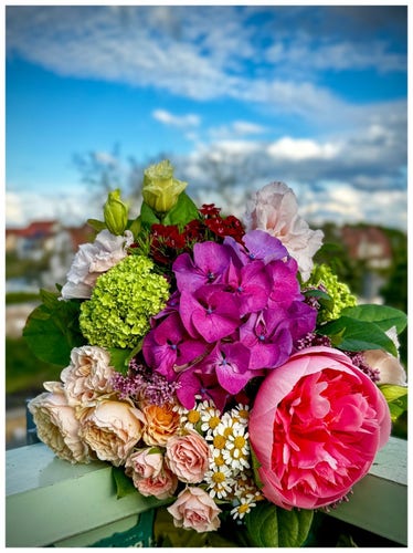 A vibrant bouquet of various flowers including roses and hydrangeas, with a blurred background of a blue sky and distant buildings.