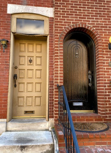 Close shot of two doors on adjoining red-brick rowhomes.  On the left is a tall rectangular door with 15 evenly spaced rectangles carved into it, in a pale buttery color.  Above that door is a square transom window, its framing a bit decayed, with a stone plinth above.  On the right is an arched door, whose bottom and top are both above those of its neighbor.  It's painted a shiny black, and carved with a series of concentric arches.  No window in, above, or near the door.  Both doors have dark brass hardware and knockers, and a blue-black iron railing divides them. The left staircase is made of white stone, while the right staircase is made of red bricks.