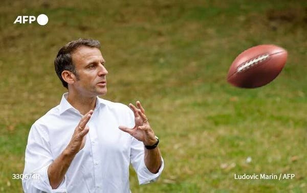 French leader Emanual Macron as if he is holding his hands out to catch the American football hurtling toward him