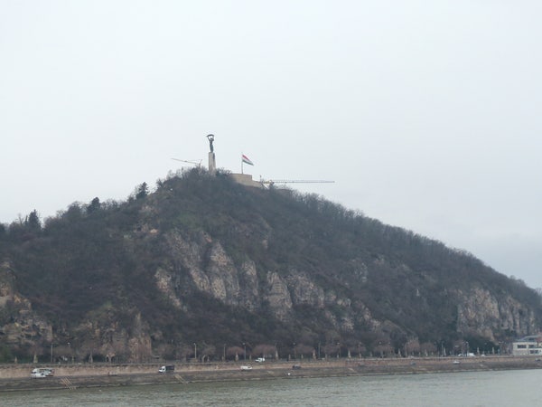 Gellért Hill visible from afar, with a hungarian flag and a tall sculpture upon it. Two construction cranes are visible, a bit less prominently