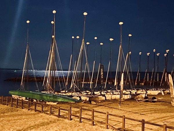 Boats neatly arranged on the beach. Between the masts the silvery light of the moon shimmers on the sea. 
