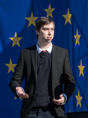 A person speaking in formal attire in front of an EU flag. At closer look, some stars in the EU flag contain icons like a CCTV camera or a smartphone.