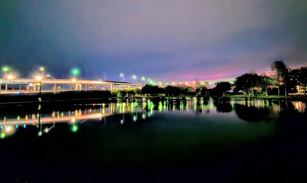 Early on a very cold morning at the edge of a large retention pond, looking across the water where shades of pink and blue appear at the horizon, beyond a well illuminated bridge with its monstrous on and off ramp structures. Glowing lights of white, yellow, and green line the roadways all reflecting upon the calm dark water below.