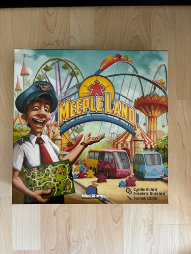 Bild eines Brettspiels namens "Meeple Land" mit einem farbenfrohen Vergnügungspark-Thema, mit einer männlichen Zeichentrickfigur, die eine offene Brettspielschachtel hält und in Richtung des Parks gestikuliert.