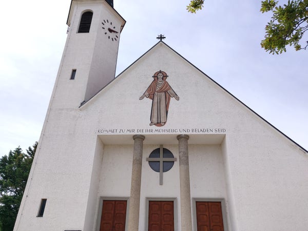 Photo of a white-painted Catholic parish church. They is a belfry with a clock face on the left. Above the three main doors there is a painted cross, above that a painting of Jesus Christ, and in between the German inscription "kommet zu mir, die ihr mühselig und beladen seid". Another cross, made of wrought iron, tops the gable of the roof.
