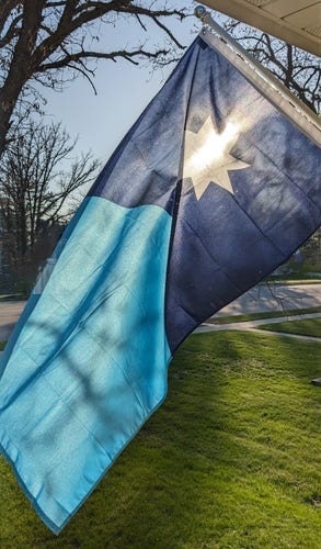 A picture of the new Minnesota State Flag flying on a white flag pole with the sun shining behind where the star is located.