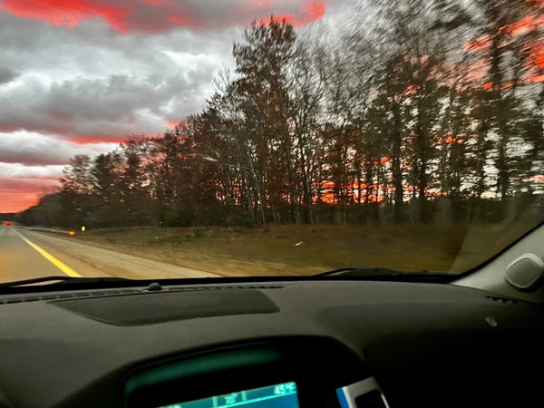 A view from inside a vehicle showing a road with a sunset and trees on the horizon. The sky has scattered clouds tinged with red and orange hues.