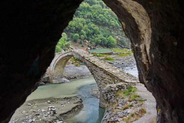 Kamienny most przerzucony nad rzeką, widziany przez naturalną skalistą ramę. Ludzie gromadzą się na moście i brzegach rzeki, otoczeni bujną zielenią.