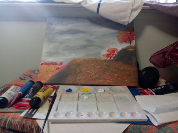 a canvas set-up atop a dresser. the canvas pictures a large and hilly summer-or-fall-time field, with distant red trees. there is a palette of paint set up in front of it, alongside brushes and paint tubes.