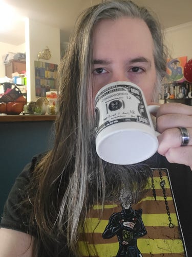 Guy with long gray hair and beard in Pinhead shirt drinks coffee from a mug with $100 on if