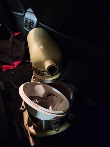 An old straw-colored bowl-lift kitchenaid sitting in the back seat of a truck with most of its parts in the bowl. It's missing the cap over the accessory port.