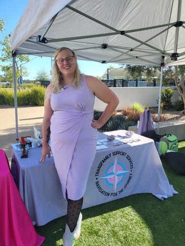 Photo of me in a lavender color dress wearing sparkly bedazzled western booties in front of our booth for TransFamily Support Services