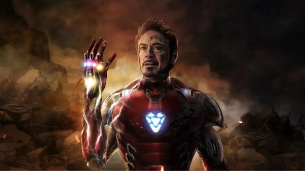 Illustration I found online of Iron Man the Marvel character played by Robert Downey Jr.. He’s in his robots suit, but his helmets off. He’s holding his right arm up as different color energy flows through it. He’s got a heart shaped light in the center of his chest. The background is smoky and on fire but dark.
