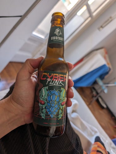 An empty brown beer bottle with a label reading CYBER PUNK. Its logo is a split-in-two Terminator-style metal skull with a cluster of hops inside of it. It was photographed in the flat of Bruce Sterling and Jasmina Tesanovic in Turin, Italy. Parts of the flat are visible in the background.