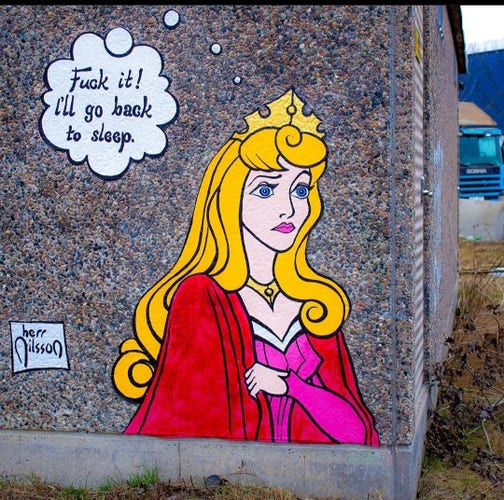 Streetartwall. On a small street wall is a mural of a very tired princess. The young lady with long blonde hair, blue eyes, crown, pink dress and red cape has a comic-like thought bubble next to her. It reads: "Fuck it! i'll go back to sleep."