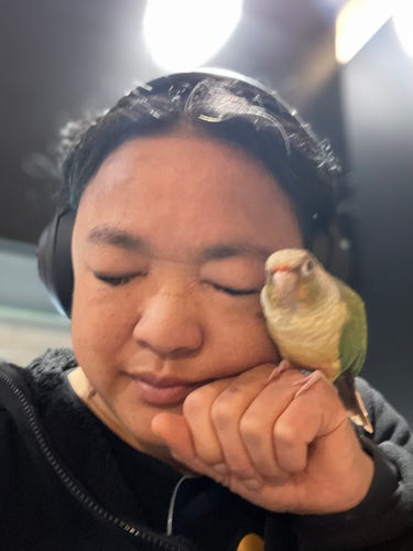 An Asian woman with a yellow and green parrot on her hand. She has her eyes closed and is clearly enjoying the snuggles
