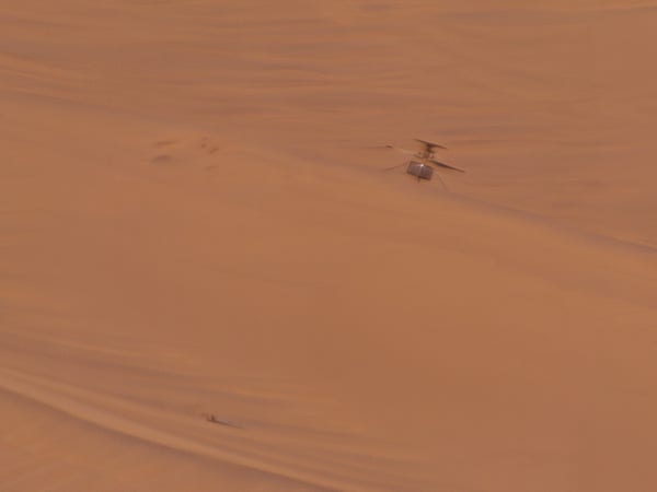 long distance view of the Ingenuity Mars Helicopter, it stands on the backside of a sand ripple and small impressions in the sand are visible to the left of it. Visible below in the image is the detached rotor blade which lies ~15m away from the heli. 