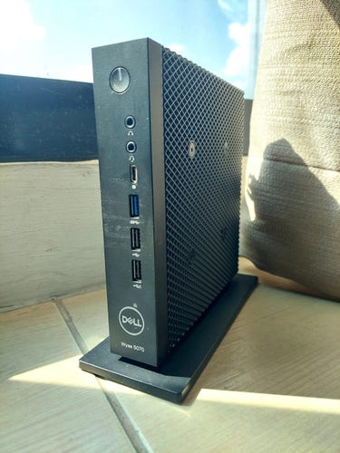 A Dell Wyse 5070 thin client computer. It's a small, slim, black PC, a vertical box about 7 inches tall and 7 inches deep, and only about 1.5 inches thick, mounted on a plastic base. You can see the power button and various ports on the narrow front panel, along with the Dell logo. The side panels have a grid-pattern surface structure that makes it look like a server computer, and probably helps with cooling the fanless system.