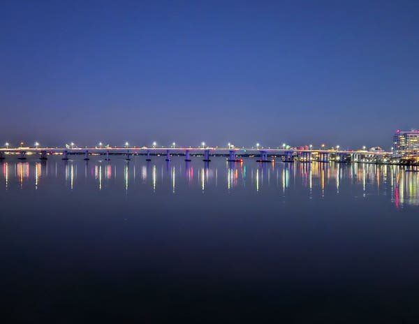 A clear, dark blue night sky over calm dark waters of a vast river where a massive interstate highway bridge spans the distance between the colorfully illuminated skylines at each shoreline. The bridge and building lights glow in the night sky, casting long reflections on the glass like water below.