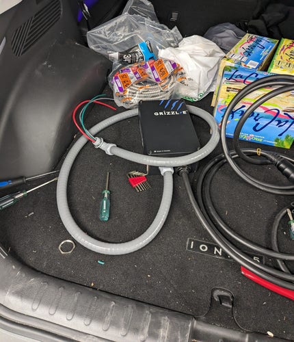 The trunk of a car filled with a wild assortment of things, including an electric vehicle charger with a grey wire whip attached to one end, MC armored cable, various tools, some LaCroix boxes, and a Menards bag.