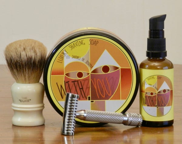 A silvertip badger shaving brush with a white handle on which "The 'Duke'" is printed in black stands next to a tub of shaving soap whose label shows a simplified version of the center of Paul Klee's painting Senecio (or, Head of a Man Goiing Senile). "Luxury Shaving Soap" and "Los Jabones de Joserra" are printed in all caps with narrow lines. "With You" is printed in large caps as if hand lettered. Next to it is a small bottle with a black squirt top and the same label. In front is a polished stainless steel double-open-comb safety razor whose handle has a spiral knurling.