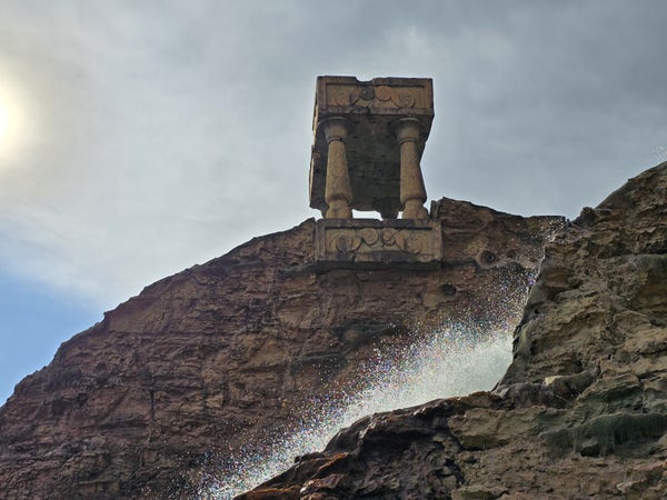 A unique structure resembling an ancient carved pillar sits atop a rugged cliff in an Italian amusement park, under a hazy sky. Sunlight catches the spray of a water feature at the cliff's base, casting a shimmering mist of rainbow-hued droplets into the air, creating a serene and mystical scene. 