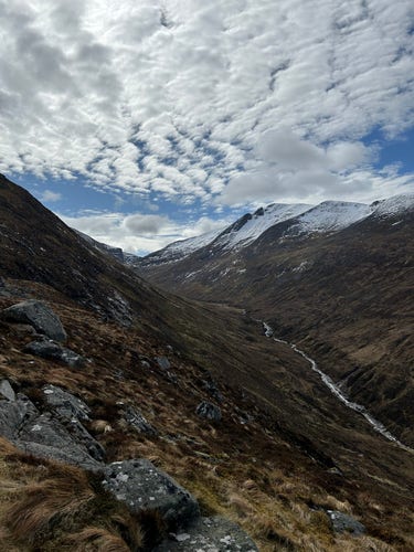 View of a snow topped Ben Nevis with clouds gathering in a blue sky and a deep valley with a silver stream