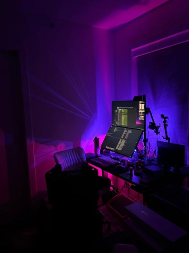 Eva's desk at home, showing two 27" 4K @ 144hz monitors, a webcam, chair, two laptops in a vertical holder on the desk surface, and various under + rear lighting from LEDs. Colors range from pink to purple and gradients in-between.