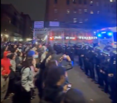 Flanks of police face hundreds of protesters in the streets of New York.  A bus can be seen in the background, which is being loaded with students.