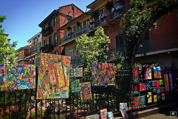 Color photo of colorful paintings hanging for sale outdoors on a black iron fence. In the background is a row of brick buildings with iron rail balconies. 