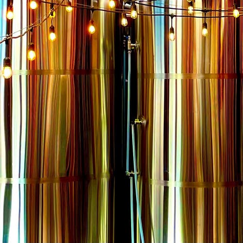 Closely cropped photo of two large mash tuns in a brewery with reflections resulting in vertical strips in shades of red, orange, and yellow. There is also a couple of strings of globe lights near the top