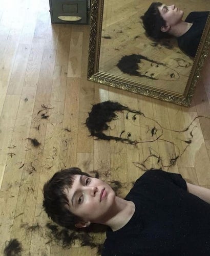 Photography. A color photo of a young woman and her cropped hair on the floor. A young woman with very short black hair lies on the wooden floor in front of a mirror and looks confidently into the camera. Next to her lies her damp and freshly cut hair, from which she has drawn a fine face with short black hair with her fingers. It looks like her. A wonderful idea.
Info: Lena Brauner is a Czech artist who makes beautiful fantasy drawings and paintings.