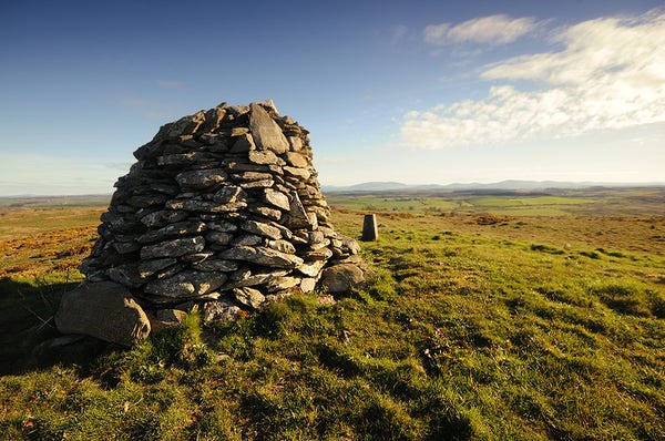 Photo of a drystone cairn and trig point on a hill top on a fine spring day, under a blue sky.