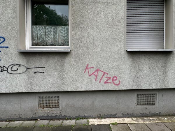 a grey pebbledash wall with two windows and some graffiti tags. One of them reads “KATze” in pink