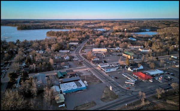 Aerial view of mainstreet in a small town. Scattered commercial businesses, surrounded by homes. There is a lake in the background.