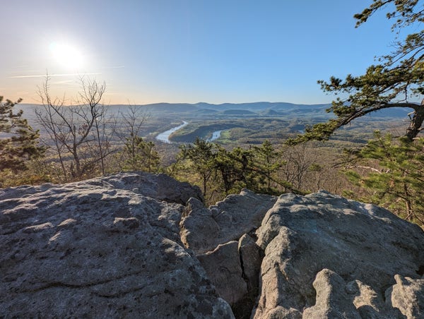 A view from the top of Little Crease mountain in the George Washington National Forest. In the background, the sun rises above a mountain ridge line on the horizon, with the winding Shenandoah River in the valley in front of it. In the foreground is an out-cropping of rocks and a few tree limbs.