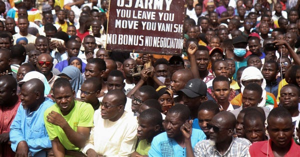Crowd of Nigerians under a brown protest sign with white stencil that says, "US army you leave, you move, you vanish. No bonus.  No negociation."