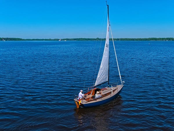 A small sailing ship on a large lake. The water is as blue as the sky. There is a forest on the shore. There are two people on board the ship. One person is waving. It is a sunny, warm day.