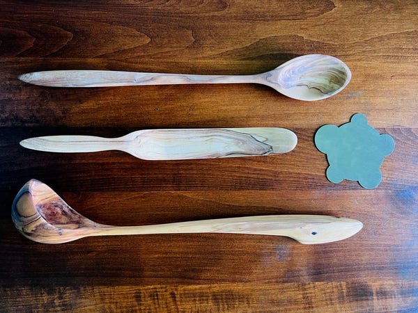 Three kitchen utensils made from olive wood: a spoon, a spurtle (which is a long, narrow, gently-curved spatula perfect for getting the last bit of mayonnaise out of the jar) and a ladle. The grain is prominent in the ladle, which has had some oil rubbed in. Also pictured: a metal cabinet scraper, which is a flat piece of metal punched out into a roughly turtle-shaped design, so that each of the legs and the head are a different circular diameter. The varying sizes make each lobe ideal for a slightly different smoothing task.