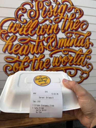 A to-go box of food from Senor Sisig. The french fries are described as being "hella spicy" on the receipt on the side of the box. In the background is a mural that says "Sisig will win the hearts and minds of the world" in a very cool and cursive font.