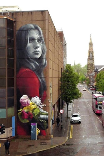 Streetartwall. A huge (16x5m) portrait of a young woman is painted on the outside wall of a brown tower block. The painting style is photorealistic and the young woman with half-length brown hair and a red sweater is depicted up to her hips. She stands slightly to one side and looks sadly at the viewer. In the lower part of the mural is a reclining bouquet of colorful flowers. (The photo shows a view of the wide main street with buses and a church in the background next to the building)