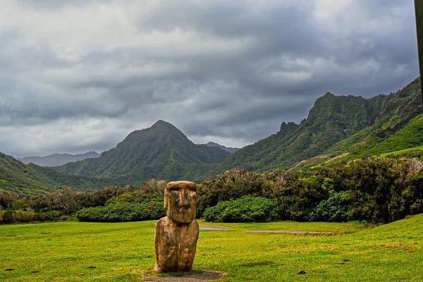 A stone statue stands in a lush green meadow with misty mountains and cloudy skies in the background.