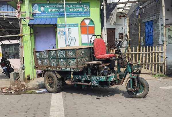 A jugaad (innovative/frugal workaround) mini-pick up truck. It has a bright red colored single seater for the driver with a cargo area at the rear. The tyres are as small as that of a scooter. The steering wheel is round in shape. On the background, you see a poster of Mamata Banerjee (the current Chief Minister of West Bengal) and a local candidate hung on a wall of tiny house. There's a small window on the house with the glasses painted in a mix of Orange, Green, and White - the colors of the Trinamool Congress (TMC) party. 