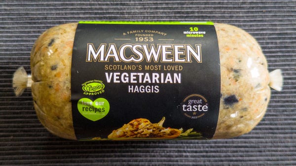 A plastic tube filled with MacSween’s vegetarian haggis. The haggis is a mix of pulses, beans, and spices, and is yellow-brown in this uncooked state. The label is mostly black with white and white writing and green accents. The background is a ridged grey table place setting.