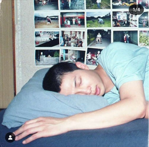 RM of BTS sleeping, with his head on a grey pillow. Behind him are photos arranged in a grid on the wall.